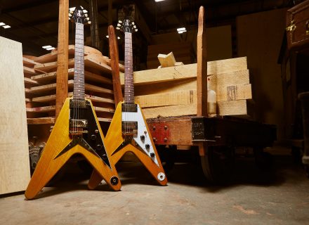 Two Gibson Custom Shop 1958 Korina Flying V Reissues (one in Black and White) behind the backdrop of a Gibson warehouse.