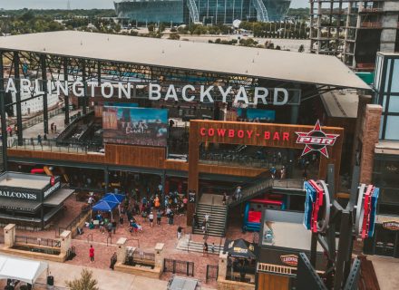 A large two-story open-air venue with a large sign above it saying Arlington Backyard. Two large TVs can be seen along with a bar area called Balcones and a smaller sign reading Cowboy Bar above the second floor.