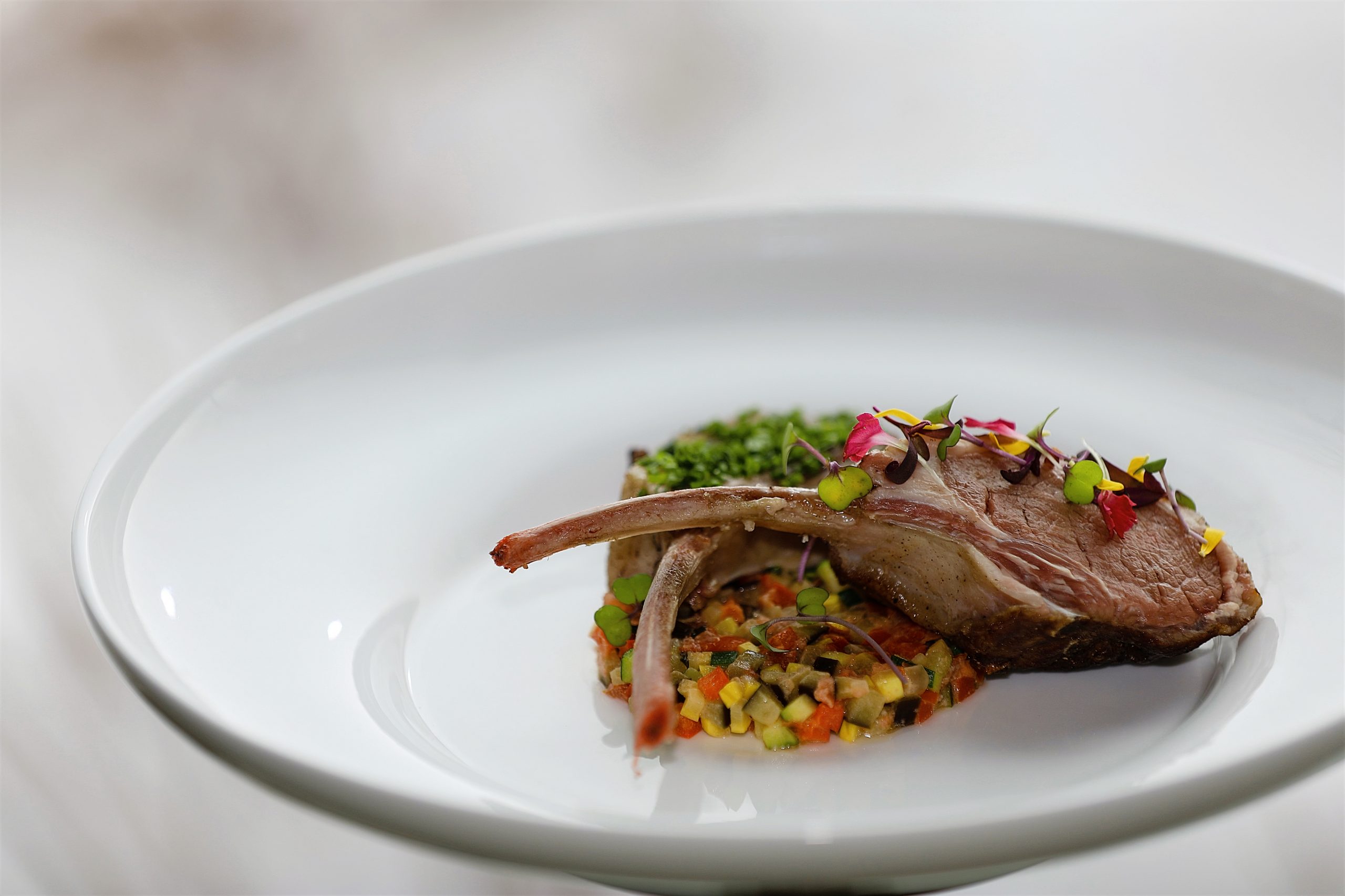 A lamb chop garnished with colorful florals sits on a bed of greenhouse ratatouille from the Brush Creek Ranch menu sits on a white plate.